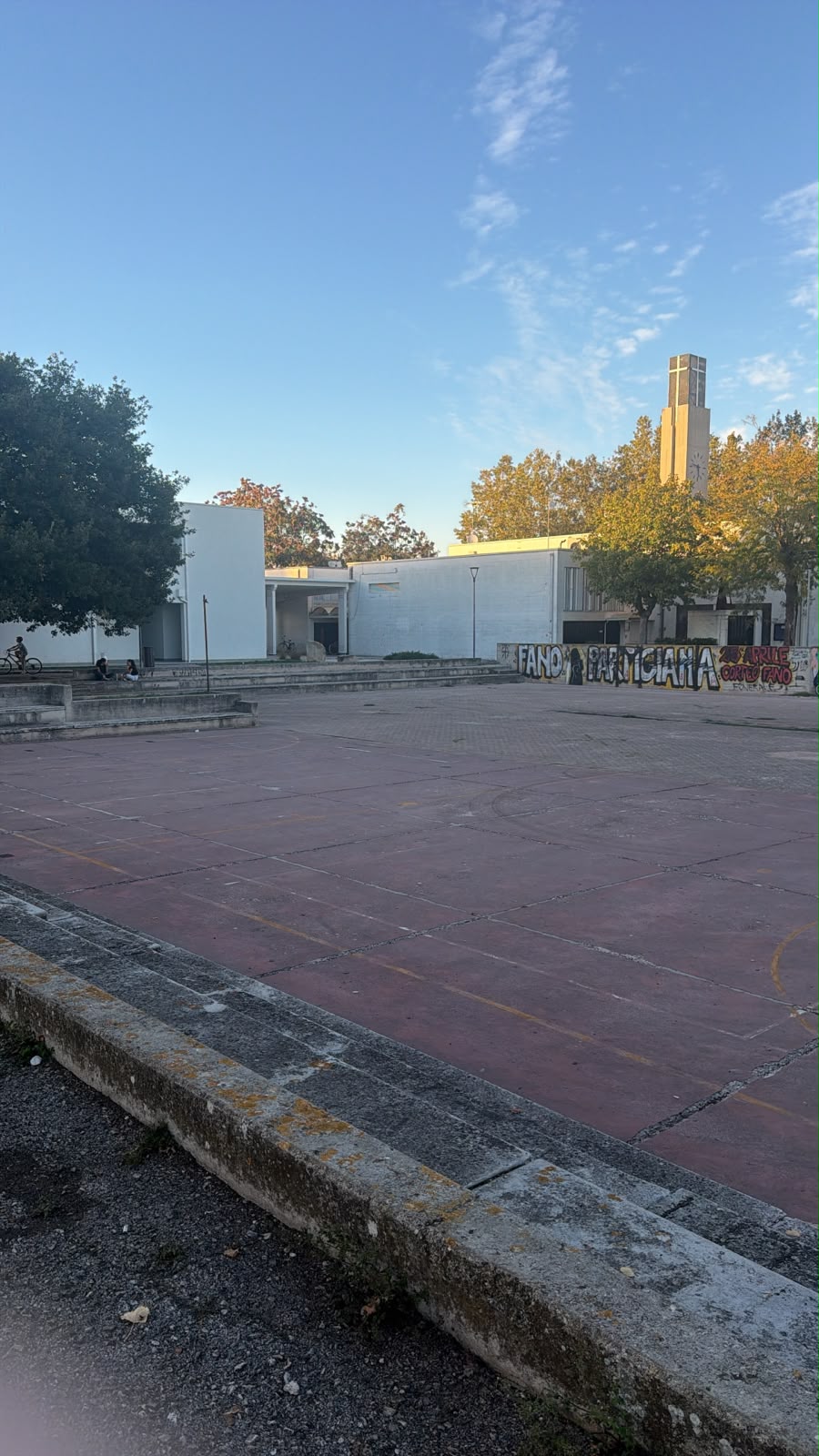 Uno spazio pavimentato del Vallato, con gradinate, alberi, edifici e una torre con croce sullo sfondo.